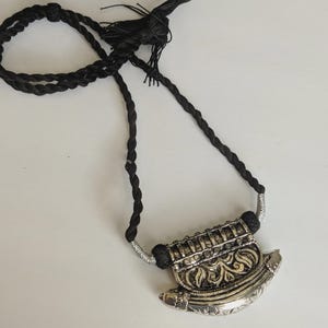 May include: A silver-toned pendant necklace with a black braided cord. The pendant has an ornate design with floral and geometric patterns. The necklace is displayed on a white surface.