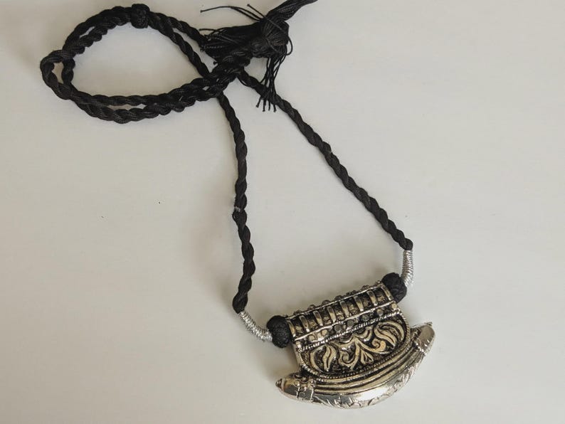 May include: A silver-toned pendant necklace with a black braided cord. The pendant has an ornate design with floral and geometric patterns. The necklace is displayed on a white surface.