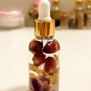 May include: A clear glass bottle with a gold cap and white dropper. The bottle contains a clear liquid with dried red flowers, gold flakes, and purple crystals. Other bottles are visible in the blurred background.