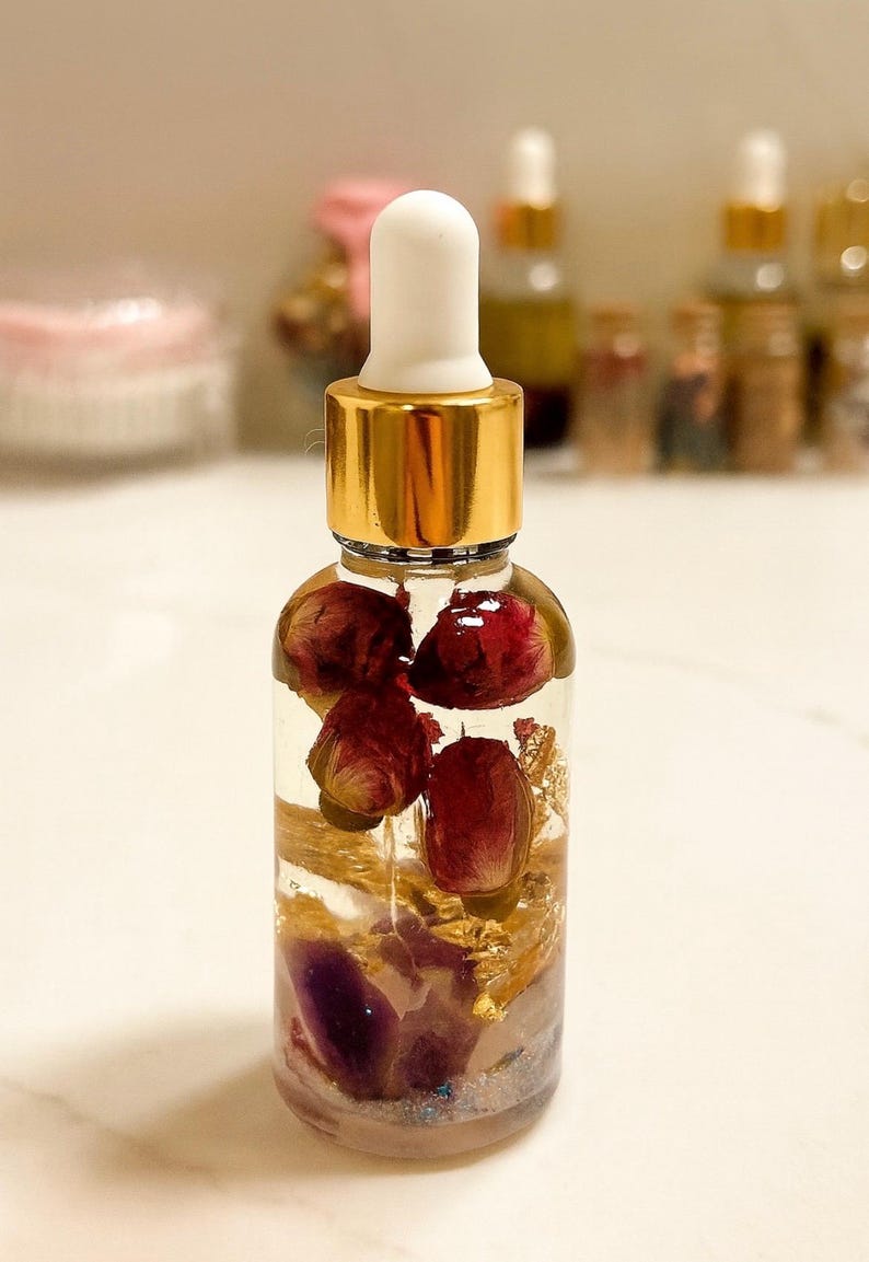 May include: A clear glass bottle with a gold cap and white dropper. The bottle contains a clear liquid with dried red flowers, gold flakes, and purple crystals. Other bottles are visible in the blurred background.