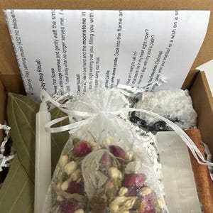 Puede incluir: Un kit de ritual espiritual con una vela blanca, una bolsita de flores secas, un cristal, una rama de canela y una hoja de laurel. El kit incluye instrucciones para un ritual, con texto visible en el papel.