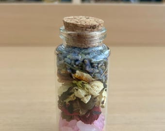 Puede incluir: Una pequeña botella de vidrio transparente llena de capas de flores secas, cristales y hierbas. La botella está rematada con un tapón de corcho. El contenido incluye lavanda, pétalos de rosa y cristales rosas. La botella mide unos 7.6 cm de alto.