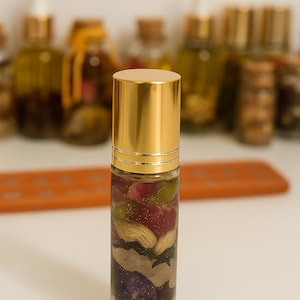 Pode incluir: Um frasco roll-on de vidro transparente com tampa dourada, preenchido com uma mistura de botânicos coloridos. O conteúdo inclui pétalas vermelhas, pedaços verdes e nozes bege. Outras garrafas estão desfocadas no fundo.