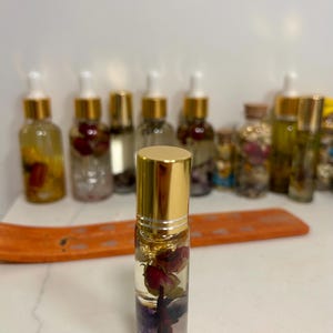 May include: A clear glass roll-on bottle with a gold cap, filled with a clear liquid and dried flowers. The bottle contains rose petals and gold flakes. Other bottles with gold caps and clear liquid are in the background.