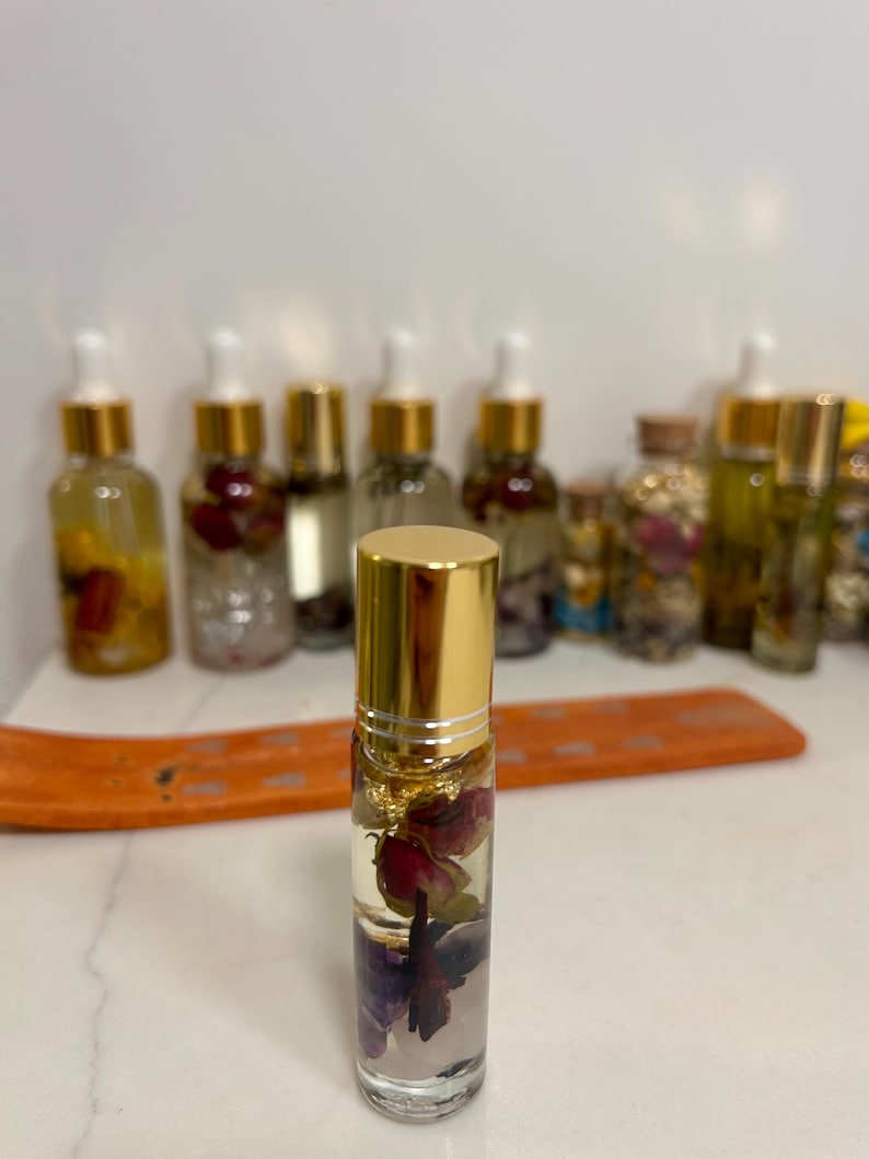 May include: A clear glass roll-on bottle with a gold cap, filled with a clear liquid and dried flowers. The bottle contains rose petals and gold flakes. Other bottles with gold caps and clear liquid are in the background.
