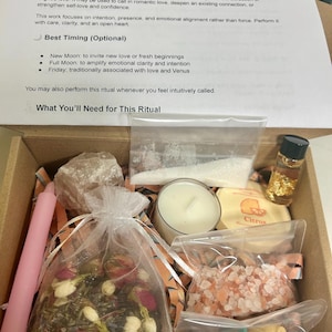 Könnte beinhalten: Eine Box mit einem Liebes- und Anziehungszauber-Set. Das Set enthält einen Beutel mit Kräutern, rosa Kerzen, eine Kerze, Kristalle, Badesalz, eine kleine Flasche und Anweisungen. Die Anweisungen sind auf weißem Papier gedruckt.