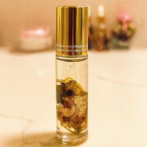 May include: A clear glass roller bottle with a gold cap, filled with a clear liquid and gold flakes. The bottle is standing upright on a light-colored surface. The background is blurred, with hints of other bottles and a pink object.