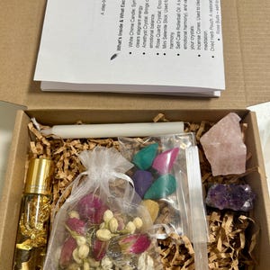 Puede incluir: Una caja de cartón abierta llena de varios artículos espirituales. Contiene un aceite en roll-on, una vela blanca, cristales, conos de incienso, flores secas y una pequeña bolsa de hierbas. La caja también incluye una lista de los artículos y sus significados.