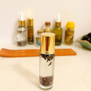 May include: A clear glass essential oil roller bottle with a gold cap, filled with dried herbs and purple flower petals. Several other bottles are in the background. An orange wooden tray is in the background.
