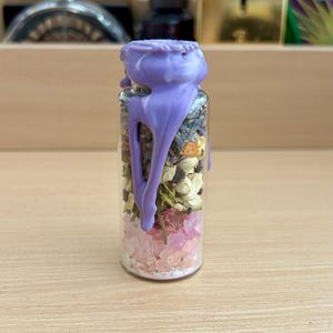 May include: A small glass bottle filled with dried flowers, pink crystals, and white granules. The bottle is sealed with a purple wax, which drips down the sides. The background is blurred, with other bottles visible.