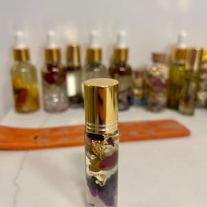 May include: A clear glass essential oil roller bottle with a gold cap, filled with dried flowers, gold flakes, and crystals. Other similar bottles are in the background. The bottle is on a white surface.