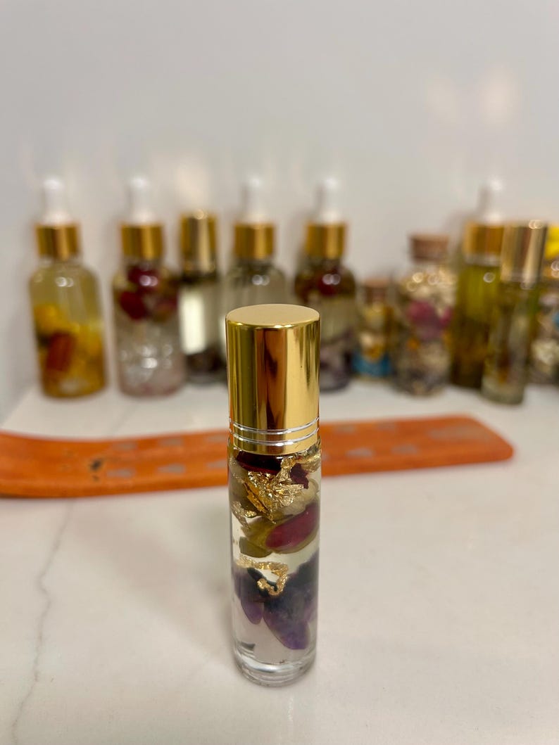 May include: A clear glass essential oil roller bottle with a gold cap, filled with dried flowers, gold flakes, and crystals. Other similar bottles are in the background. The bottle is on a white surface.