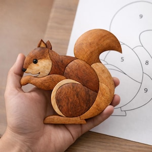 Puede incluir: Un rompecabezas de madera con forma de ardilla, compuesto por piezas de diferentes tonos marrones. Un diagrama es visible en el fondo. La ardilla tiene una cola curvada y se sostiene en una mano.