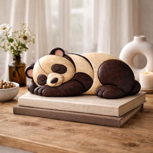 May include: A wooden panda sculpture in a sleeping pose, with dark brown and light wood tones. The panda rests on a stack of two books. The sculpture is on a wooden table, with a vase of flowers and a candle in the background.