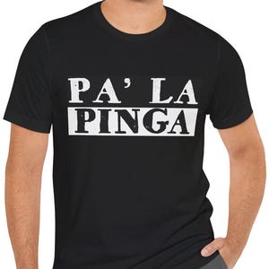 May include: Black t-shirt with the white text "PA' LA PINGA" in a bold, blocky font. The shirt is a crew neck style and is being worn by a person. The person is wearing gray pants.