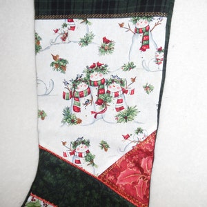 May include: A Christmas stocking with a festive snowman design. The main fabric features snowmen with red and green scarves, surrounded by birds and holly. The top and bottom sections are green and red patterned fabrics.