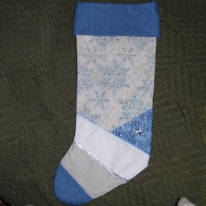 May include: A handmade Christmas stocking with a blue denim cuff and toe. The body features a snowflake pattern on a cream background, with sections of white, silver, and blue fabric. A silver star embellishes the blue section.