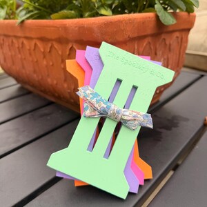 May include: A stack of colorful, slotted craft tools in green, purple, pink, and orange, with a floral bow. The text "The Spoolery & Co." is printed on the green tool. A terracotta pot with green plants is in the background.