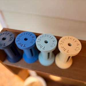 May include: Four wooden spools in shades of blue and tan are arranged on a wooden shelf. Each spool has the text "The Spoolery & Co." printed on the top. The spools are painted in navy, blue, light blue, and tan.