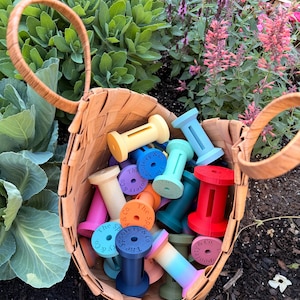 May include: A woven basket overflowing with colorful wooden spools. The spools are in various shades including yellow, blue, pink, and red. Each spool has the text "The Spoolery & Co." printed on it. The basket is set against a backdrop of green foliage and flowers.