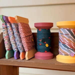 May include: A wooden shelf displays colorful spools of ribbon. The spools have various designs, including floral and striped patterns. The spools are in shades of green, purple, orange, and yellow. The shelf is made of wood and has a natural finish.