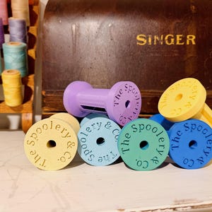 May include: A collection of colorful thread spool holders in various pastel shades, including yellow, blue, green, and purple. Each holder is round with a central hole and the text "The Spoolery & Co." The background includes spools of thread and a vintage Singer sewing machine.