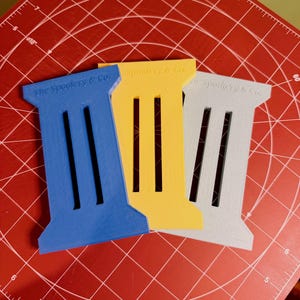 May include: Three rectangular, stylized objects in blue, yellow, and gray, each with three vertical slots and the text "The Spookery & Co." on top. They are arranged on a red cutting mat with grid lines.