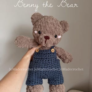 Può includere: Un orsacchiotto all'uncinetto marrone fatto a mano chiamato "Benny the Bear" con una tuta blu con bottoni di legno. L'orso ha occhi neri e accenti rosa. Il testo "DIGITAL PATTERN" è in basso.
