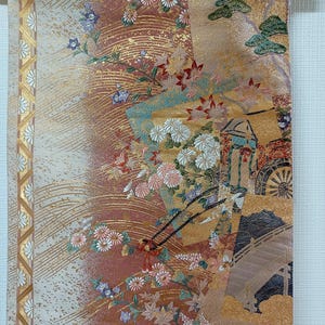 May include: A decorative Japanese scroll featuring a detailed design. The artwork showcases a blend of colors, including gold, red, and cream, with floral and landscape elements. The scroll is suspended from a wooden rod with a natural fiber cord.