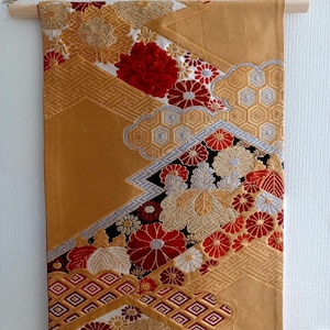 May include: A decorative wall hanging with a gold background and intricate floral patterns in red, gold, and white. The design includes geometric shapes and is suspended from a wooden rod.