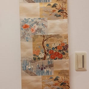 May include: A long, rectangular wall hanging with a beige background and multiple panels featuring floral and landscape designs. The artwork includes irises, bamboo, and flowering trees, with a wooden rod at the bottom and top for hanging.