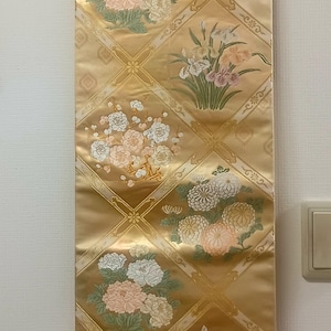 May include: A decorative wall hanging with a gold background and floral patterns. The design features various flowers in shades of pink, white, and green, arranged in diamond-shaped sections. The hanging is suspended from a wooden rod.