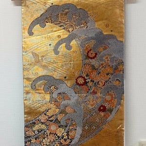 May include: A decorative wall hanging featuring a stylized wave design. The artwork uses gold, gray, and floral patterns. A bird is depicted in the background. The piece is mounted on a wooden rod with a hanging cord.