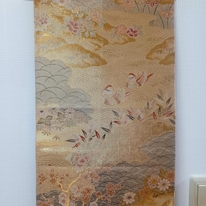 May include: A decorative Japanese wall hanging with a floral and bird design. The fabric features a gold, beige, and gray color palette with detailed illustrations of flowers, birds, and landscapes. The hanging is suspended from a wooden rod.