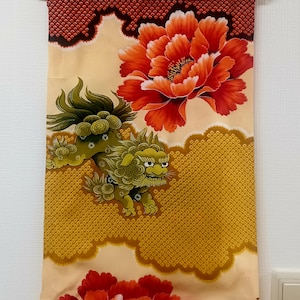 May include: A decorative wall hanging featuring a vibrant design. The artwork showcases a large, red peony flower and a green lion-like creature. The background has a gold and red pattern. The top border is a dark red pattern.