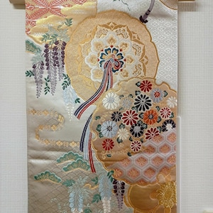 May include: A decorative Japanese wall hanging with a wooden rod. The textile features a detailed floral design in gold, cream, and pastel colors. Intricate patterns and hanging tassels add to the traditional aesthetic.