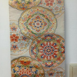 May include: A decorative textile with a repeating pattern of large, ornate floral medallions in shades of gold, orange, blue, and pink. The background is a light cream color. The textile is hanging from a wooden rod.