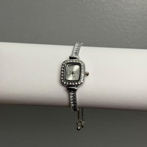 May include: A silver-toned wristwatch with a rectangular face and a bezel encrusted with small, clear crystals. The watch has a silver band and a quartz movement. The watch is displayed on a white surface against a gray background.