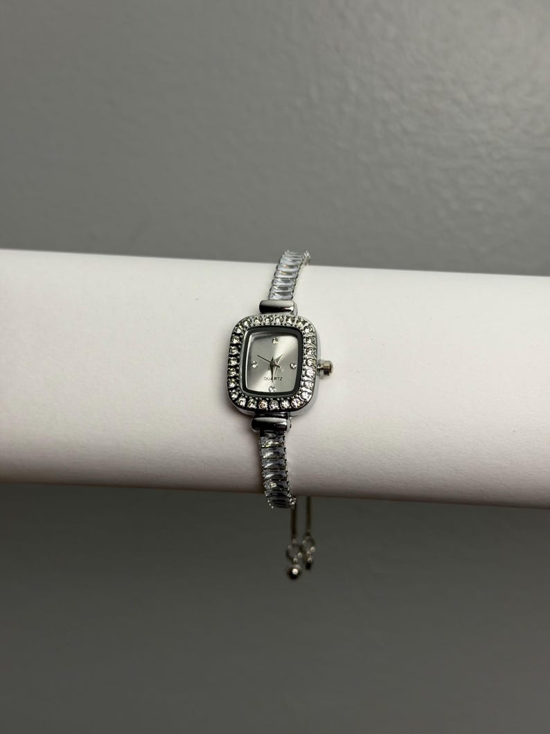 May include: A silver-toned wristwatch with a rectangular face and a bezel encrusted with small, clear crystals. The watch has a silver band and a quartz movement. The watch is displayed on a white surface against a gray background.