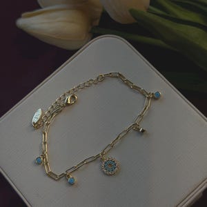 May include: Gold-toned chain bracelet with turquoise accents and a circular charm with a turquoise center and clear stones. The bracelet is on a white surface with white flowers in the background.
