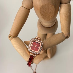 Puede incluir: Un reloj de pulsera de oro rosa con una esfera roja cuadrada y una correa de piedras rectangulares rojas. La esfera del reloj est&aacute; rodeada por un borde de peque&ntilde;os cristales transparentes. El reloj lo sostiene un maniqu&iacute; de madera.