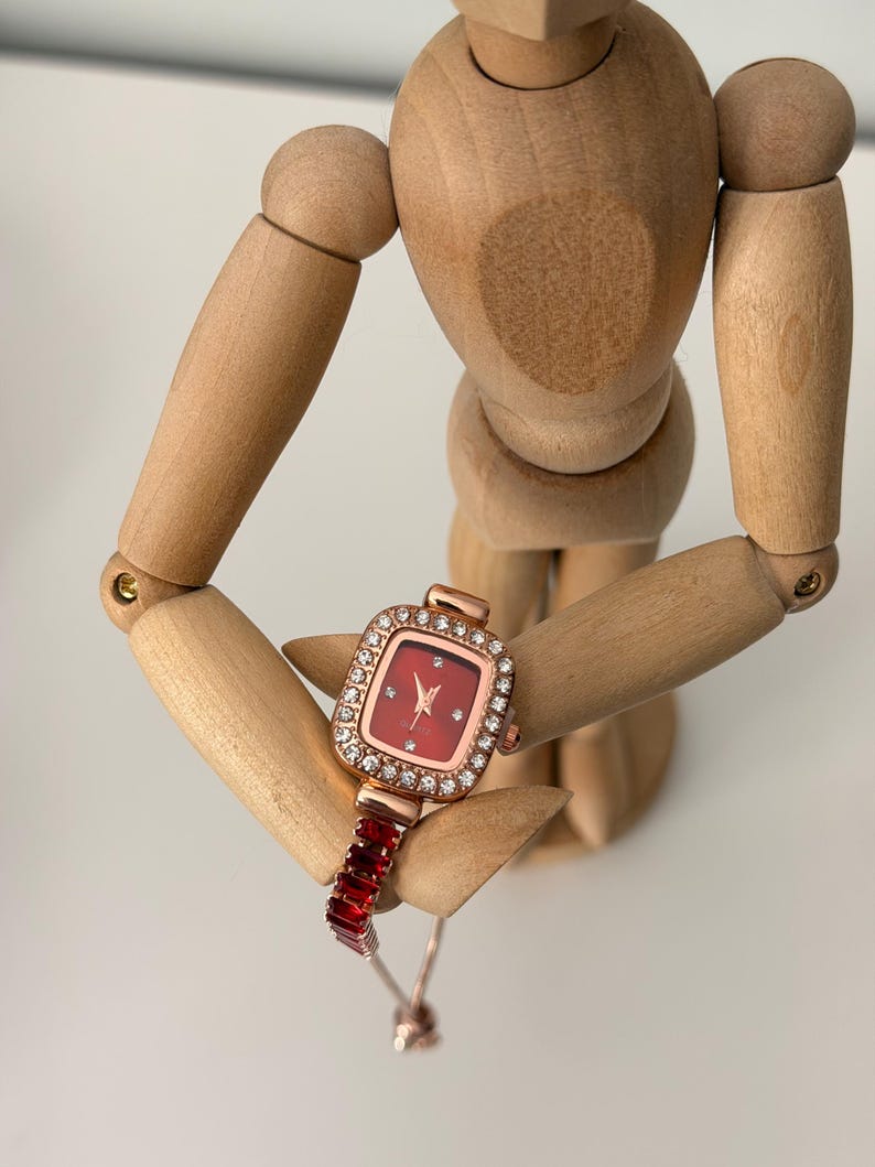 Puede incluir: Un reloj de pulsera de oro rosa con una esfera roja cuadrada y una correa de piedras rectangulares rojas. La esfera del reloj est&aacute; rodeada por un borde de peque&ntilde;os cristales transparentes. El reloj lo sostiene un maniqu&iacute; de madera.