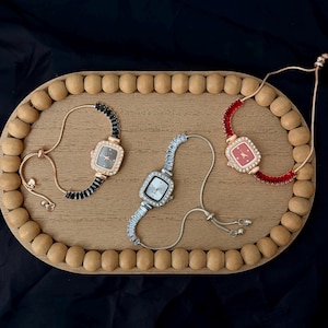 May include: Three square-faced bracelet watches on a wooden tray. One watch has a black face and black beaded band, another has a silver face and silver band, and the third has a red face and red band. The tray is oval with wooden beads around the edge.