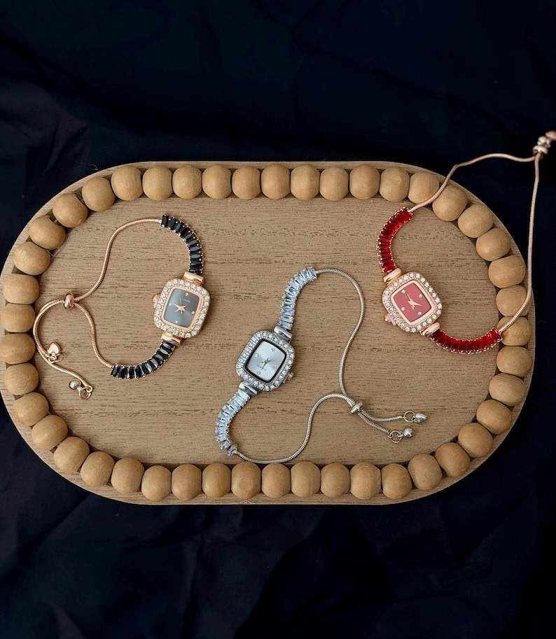 May include: Three square-faced bracelet watches on a wooden tray. One watch has a black face and black beaded band, another has a silver face and silver band, and the third has a red face and red band. The tray is oval with wooden beads around the edge.