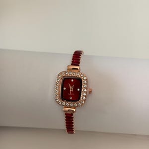 Puede incluir: Un reloj de pulsera de oro rosa con una esfera roja rectangular y un bisel incrustado con cristales transparentes. La correa del reloj es de color rojo a juego. El reloj es de movimiento de cuarzo.