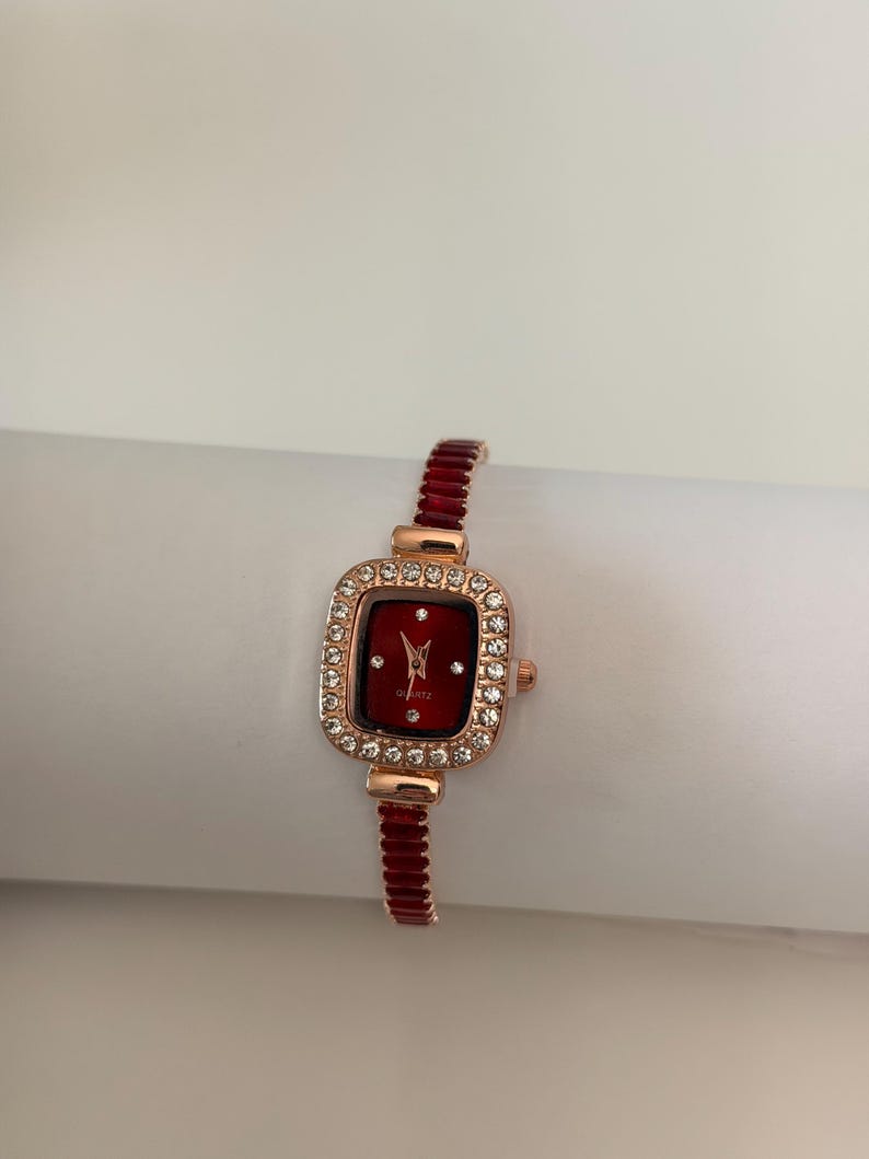 Puede incluir: Un reloj de pulsera de oro rosa con una esfera roja rectangular y un bisel incrustado con cristales transparentes. La correa del reloj es de color rojo a juego. El reloj es de movimiento de cuarzo.
