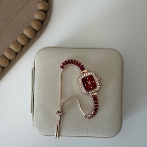 Puede incluir: Un reloj de oro rosa con una esfera cuadrada roja y un bisel incrustado con peque&ntilde;os cristales transparentes. La correa del reloj es roja con un dise&ntilde;o similar a una cadena. El reloj descansa sobre una superficie beige claro texturizada.