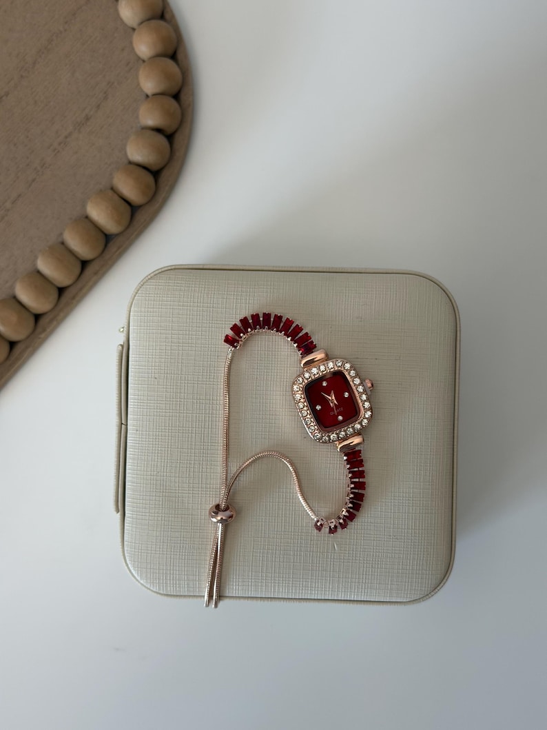 Puede incluir: Un reloj de oro rosa con una esfera cuadrada roja y un bisel incrustado con peque&ntilde;os cristales transparentes. La correa del reloj es roja con un dise&ntilde;o similar a una cadena. El reloj descansa sobre una superficie beige claro texturizada.