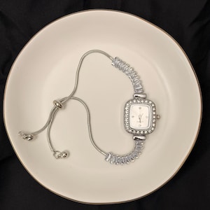 May include: A silver-toned watch with a square face, surrounded by small, clear stones. The watch is on a white plate with a gold rim, set against a black background. The watch band is a delicate chain with a sliding clasp.