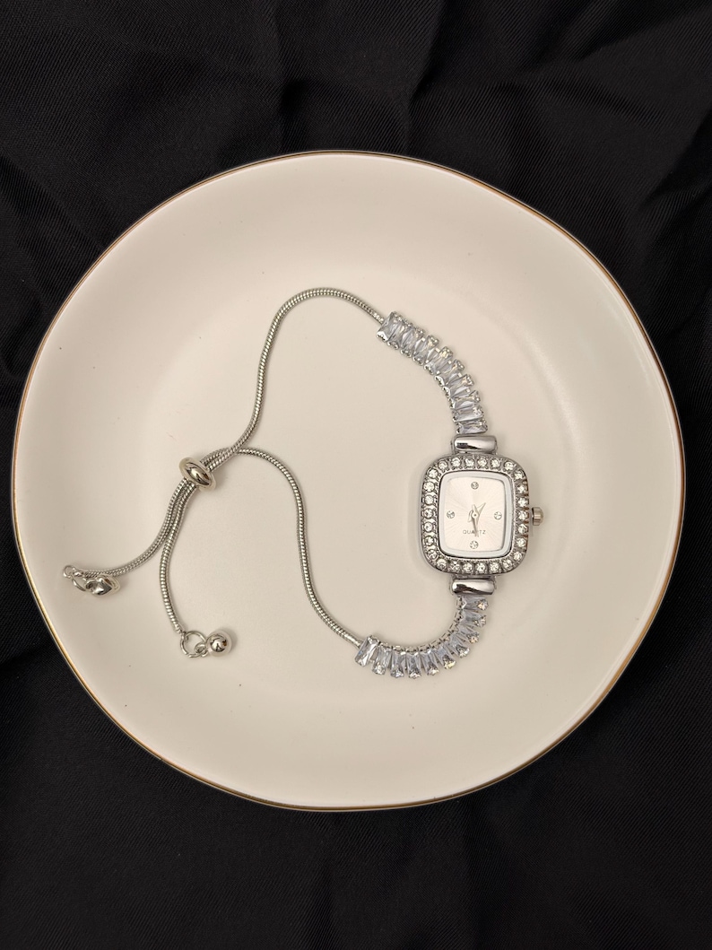 May include: A silver-toned watch with a square face, surrounded by small, clear stones. The watch is on a white plate with a gold rim, set against a black background. The watch band is a delicate chain with a sliding clasp.
